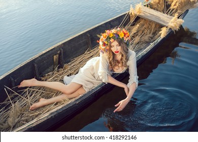 Young sexy woman on boat at sunset. The girl has a flower wreath on her head, relaxing and sailing on river. Beautiful body and face. Fantasy art photography. Concept of female beauty, rest in the