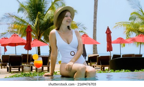 Young sexy woman in straw hat sit on edge of outdoor swimming pool in luxury hotel. Attractive girl in white swimwear smiles enjoy relax with orange cocktail drink in resort. Tropical summer vacation.