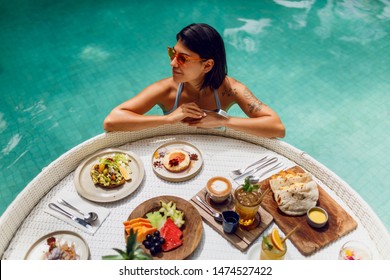 Young  Sexy Woman With Tattoo  In A Bathing Suit Having Breakfast In A Private Pool.Girl Relaxing In The Pool Drinking Coffee  And Eating Fruits. Fruit Plate, Smoothie Bowl By The Hotel Pool.