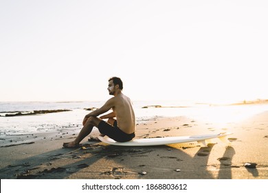 Young Shirtless Male Surfer In Shorts With Sunblock On Face Sitting On Surfing Board On Empty Sandy Seashore On Sunny Evening