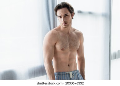 young shirtless man looking at camera near window at home