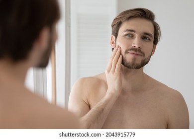 Young shirtless man looking in mirror touch his face after shave applied aftershave cream enjoy smooth, flawless, soft skin. Grooming, morning routine, ad of skincare cosmetic products for men concept