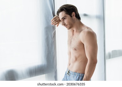young shirtless man looking through window at home