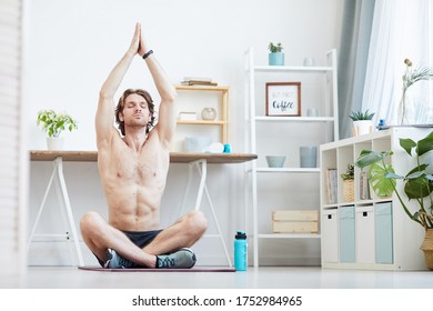 Young shirtless man sitting on exercise mat and doing breathing exercises in the living room - Powered by Shutterstock