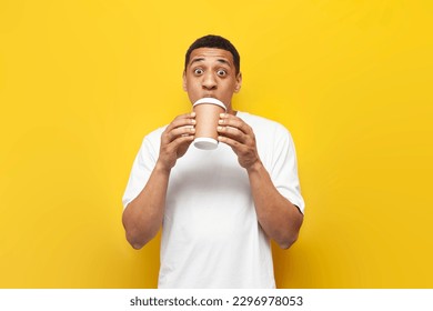 young shocked african american guy in white t-shirt drinks coffee in paper glass on yellow isolated background and shows surprise - Powered by Shutterstock