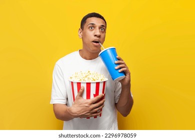 young shocked african american guy with popcorn watching movie and drinking soda on yellow isolated background, male cinema viewer is surprised