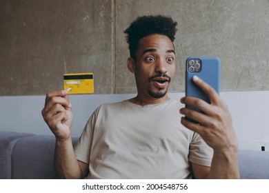 Young Shocked African American Man In Beige T-shirt Sit On Grey Sofa Indoors Apartment Hold Mobile Cell Phone Credit Bank Card Shopping Online Order Delivery Booking Tour Rest On Weekends Stay At Home