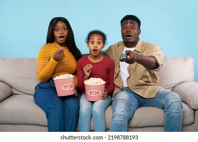 Young Shocked Black Parents And Their Daughter Watching Movie On TV And Eating Popcorn, Man Pointing At Camera With Remote Control, Sitting On Couch Over Blue Studio Background