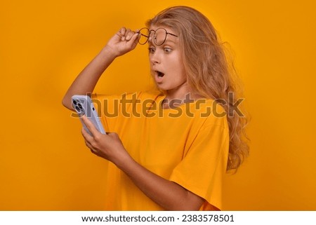 Young shocked Caucasian woman zoomer with mobile phone in hands lifts glasses from eyes and opens mouth after receiving lucrative offer to order groceries with home delivery stands in yellow studio.