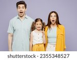 Young shocked happy parents mom dad with child kid daughter girl 6 years old wear blue yellow casual clothes looking camera with opened mouth isolated on plain purple background. Family day concept