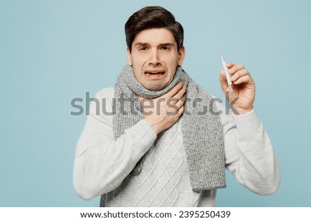 Young shocked ill sick man wear gray sweater scarf hold in hand thermometer isolated on plain blue background studio portrait. Healthy lifestyle disease virus treatment cold season recovery concept