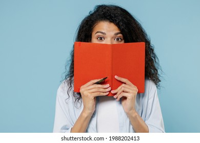 Young shocked puzzled nerd african american girl teen student in denim clothes cover mouth hiding with book notebook isolated on blue background Education in high school university college concept.