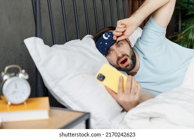 Young Shocked Sad Man Wear Blue T-shirt Lying In Bed Hold Look At Smartphone While Alarm Ringing Rest Spend Time In Bedroom Lounge Home In Own Room House Wake Up Dream. Bad Mood Night Bedtime Concept