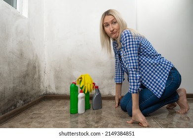 Young Shocked, Surprised Blonde Woman In Blue Jeans And A Shirt Is Squatting With Cleansers Near A Wall With Black Mold. Cleaning Concept