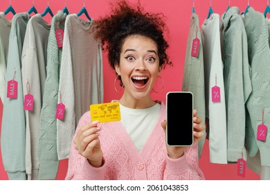 Young Shocked Woman In Sweater Stand Near Clothes Rack With Tag Sale In Store Hold Mobile Cell Phone Blank Screen Workspace Area Credit Bank Card Shop Online Isolated On Plain Pink Background Studio