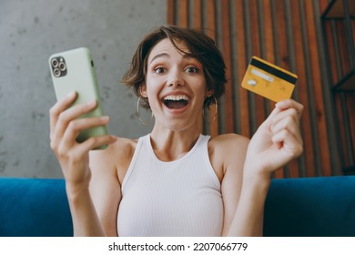 Young Shocked Woman Wear White Tank Shirt Using Mobile Cell Phone Credit Bank Card Shopping Online Booking Tour Sit On Blue Sofa Stay Home Flat Rest Relax Spend Free Spare Time In Living Room Indoors