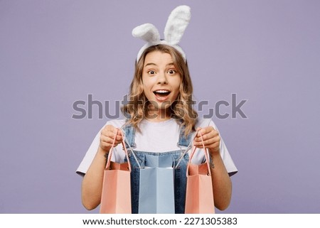 Young shocked woman wears casual clothes bunny rabbit ears hold paper package bags after shopping isolated on plain light pastel purple background studio portrait. Happy Easter sale buy day concept