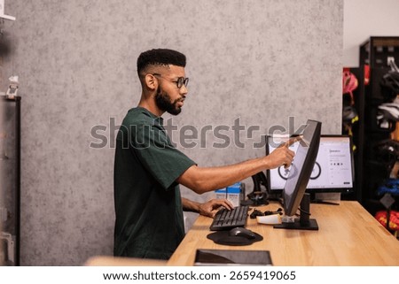 Young shop assistant pointing at computer screen while showing bike parts to customer in modern bike shop, providing professional advice and assistance