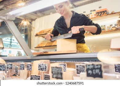 Young Shop Clerk In Deli Cutting Cheese At The Counter With A Knife