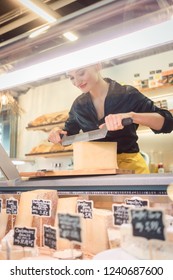 Young Shop Clerk In Deli Cutting Cheese At The Counter With A Knife