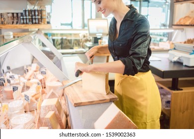 Young Shop Clerk In Deli Cutting Cheese At The Counter With A Knife