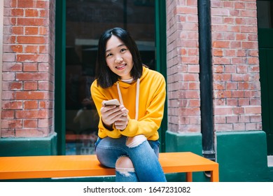 Young Short Haired Asian Woman Wearing Yellow Hoodie And Ripped Jeans With Phone In Hands Sitting On Bench Smiling At Camera