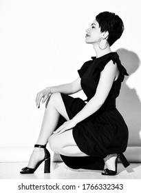 Young short haired brunette woman in elegant black dress, shoes and earrings sitting on floor and smiling over white background, side view. Casual elegance concept
