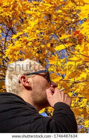 A young short-haired blond guy wearing sun glasses and a black jacket smoking in the autumn park.