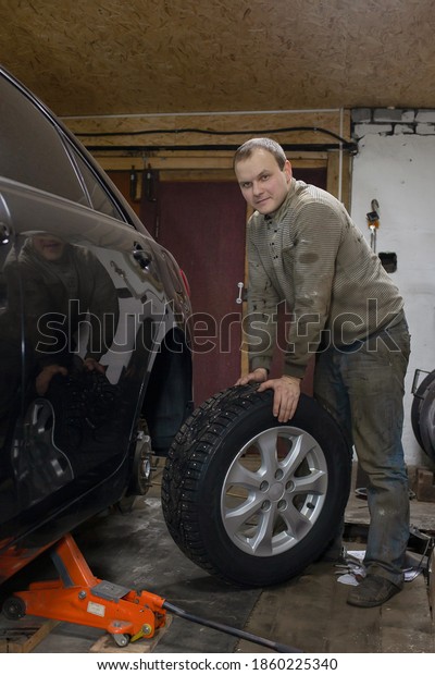 young short-haired man in\
dirty clothes rolls the wheel towards the black car which stands on\
the jack.