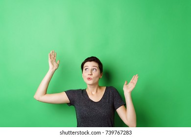 Young short-haired woman standing with hands up against chroma key green screen with copy space, looking up with whistling surprise grimace, as if she looking at invisible objects - Powered by Shutterstock