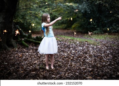 A young shy child girl with dark blond hair wearing a white tutu dress standing in a dark autumn forest surrounded by fairy sparkles pointing at one sparkle bare feet on a leave bed