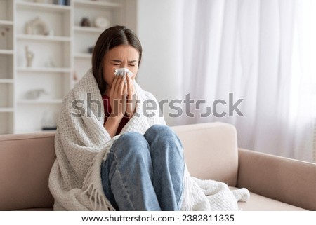 Young Sick Asian Woman Blowing Nose In Paper Tissue While Sitting On Couch At Home Covered In Blanket, Portrait Of Ill Korean Female Suffering Runny Nose, Feeling Unwell, Having Seasonal Flu