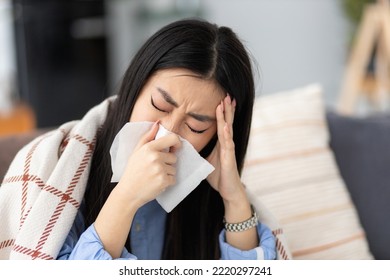 Young sick Asian woman sits on the sofa blowing her nose. Runny nose, cold or flu disease concept - Shutterstock ID 2220297241