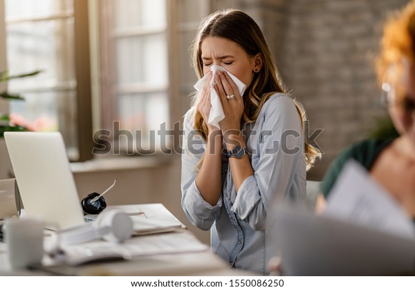 Young sick businesswoman sneezing in a tissue\
while working in the office.\
