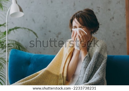 Young sick ill sad woman wears white wrapped in plaid sneezing blow nose use tissue sit on blue sofa couch stay at home hotel flat spend time in living room indoors grey wall People healthcare concept