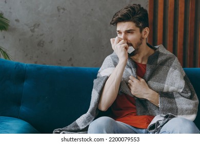 Young Sick Ill Unhappy European Cold Man Wear Red T-shirt Wrapped In Plaid Sneezing Sit On Blue Sofa Couch Stay At Home Hotel Flat Spend Time In Living Room Indoors Grey Wall. People Health Concept