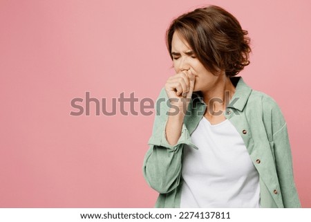 Young sick ill woman 20s she wearing green shirt white t-shirt cough hold hand near mouth isolated on plain pastel light pink background studio portrait. People healthcare pandemic lifestyle concept