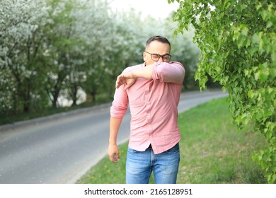 Young Sick Man Covering His Nose And Mouth With His Hand While Sneezing In The Park. Sick Infected Patient Coughing In The Elbow. Virus Protection. Proper Cough In Hands