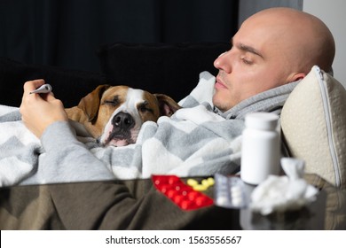 Young Sick Man Measures The Temperature Lying On The Couch With His Dog. Seasonal Cold Concept. Сoncept Of Support, Care And Friendship Between Human And His Pet.