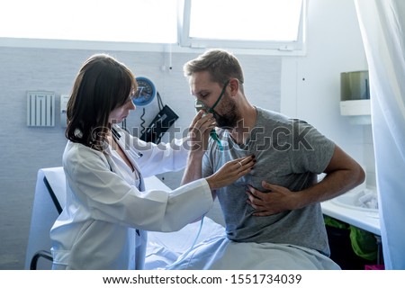 Young sick man patient with Oxygen Mask while female doctor listens his chest with stethoscope in hospital emergency room. In Smoking and respiratory diseases and anti tobacco advertising campaign.