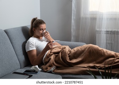 Young sick stressed woman lying at home covered with blanket checking her body temperature. Ill female have common cold and flue measuring heat with body thermometer. - Shutterstock ID 2210787451