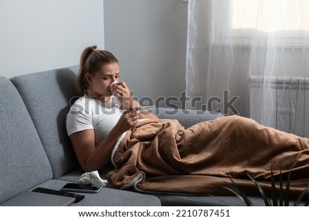 Young sick stressed woman lying at home covered with blanket checking her body temperature. Ill female have common cold and flue measuring heat with body thermometer.