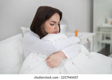 Young sick unhappy brunette woman sitting in bed at home, lady got cold, flu or coronavirus, suffering from period cramps or headache, medicine on bedside table, copy space - Shutterstock ID 2257945437