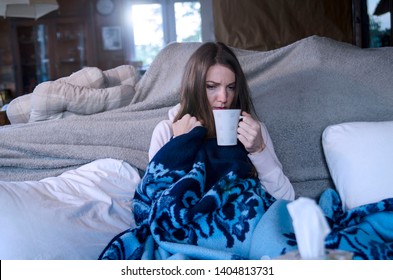 Young Sick Woman Drinking Hot Tea Sitting On Sofa Under Blanket.Miserable Woman Having Fever And Flu Virus Symptoms 