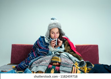 The Young Sick Woman With Flue Sitting On Sofa At Home Or Studio Covered With Knitted Warm Clothes. Illness, Influenza, Pain Concept. Relaxation At Home. Healthcare Concepts.