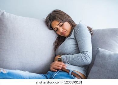 Young sick woman with hands holding pressing her crotch lower abdomen. Medical or gynecological problems, healthcare concept. Young woman suffering from abdominal pain while sitting on sofa at home
