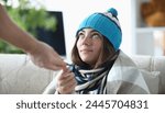 Young sick woman in hat and scarf sits under blanket on sofa at home and receives medicine. Outpatient pneumonia treatment concept.