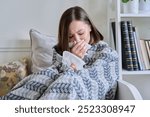 Young sick woman sitting under warm blanket sneezing coughing in handkerchief