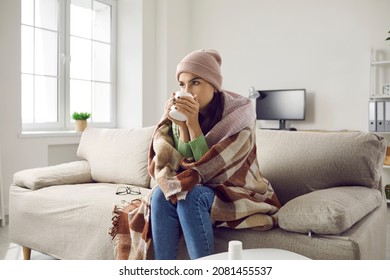 Young sick woman is treated and warmed by hot drink while sitting on sofa at home. Woman wrapped in plaid, scarf and wearing hat sitting on sofa in room with cup in hands. Home treatment concept