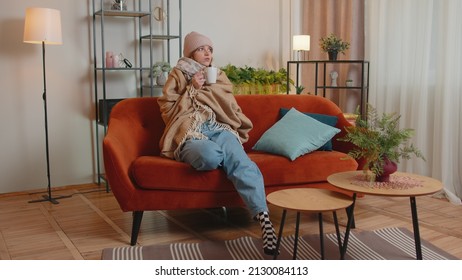 Young sick woman wear hat wrapped in plaid sit alone shivering from cold on sofa drinking hot tea in unheated apartment without heating due debt. Unhealthy female feeling discomfort try to warming up